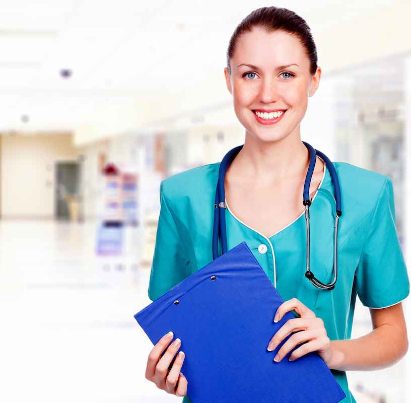 Uśmiechnięta pielęgniarka w zielonym fartuchu ze stetoskopem, trzymająca dokumentację medyczną, stojąca na korytarzu szpitalnym i patrząca w obiektyw.
